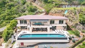 Ein modernes, zweistöckiges Haus mit Swimmingpool, überdachter Terrasse und Blick auf eine Hügelkuppe.