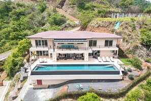 Ein modernes, zweistöckiges Haus mit Swimmingpool, überdachter Terrasse und Blick auf eine Hügelkuppe.