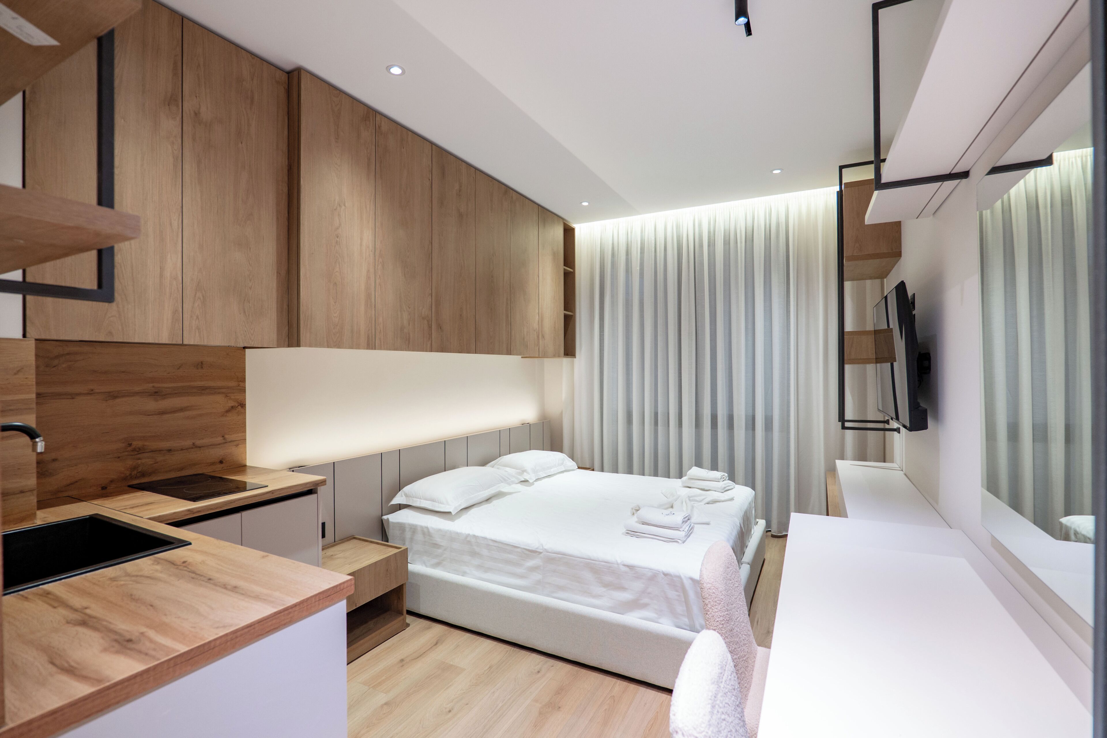 A modern hotel room with a wooden accent wall, a large bed, a bedside table, a built-in TV, and a large window with curtains.