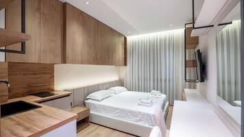A modern hotel room with a wooden accent wall, a large bed, a bedside table, a built-in TV, and a large window with curtains.