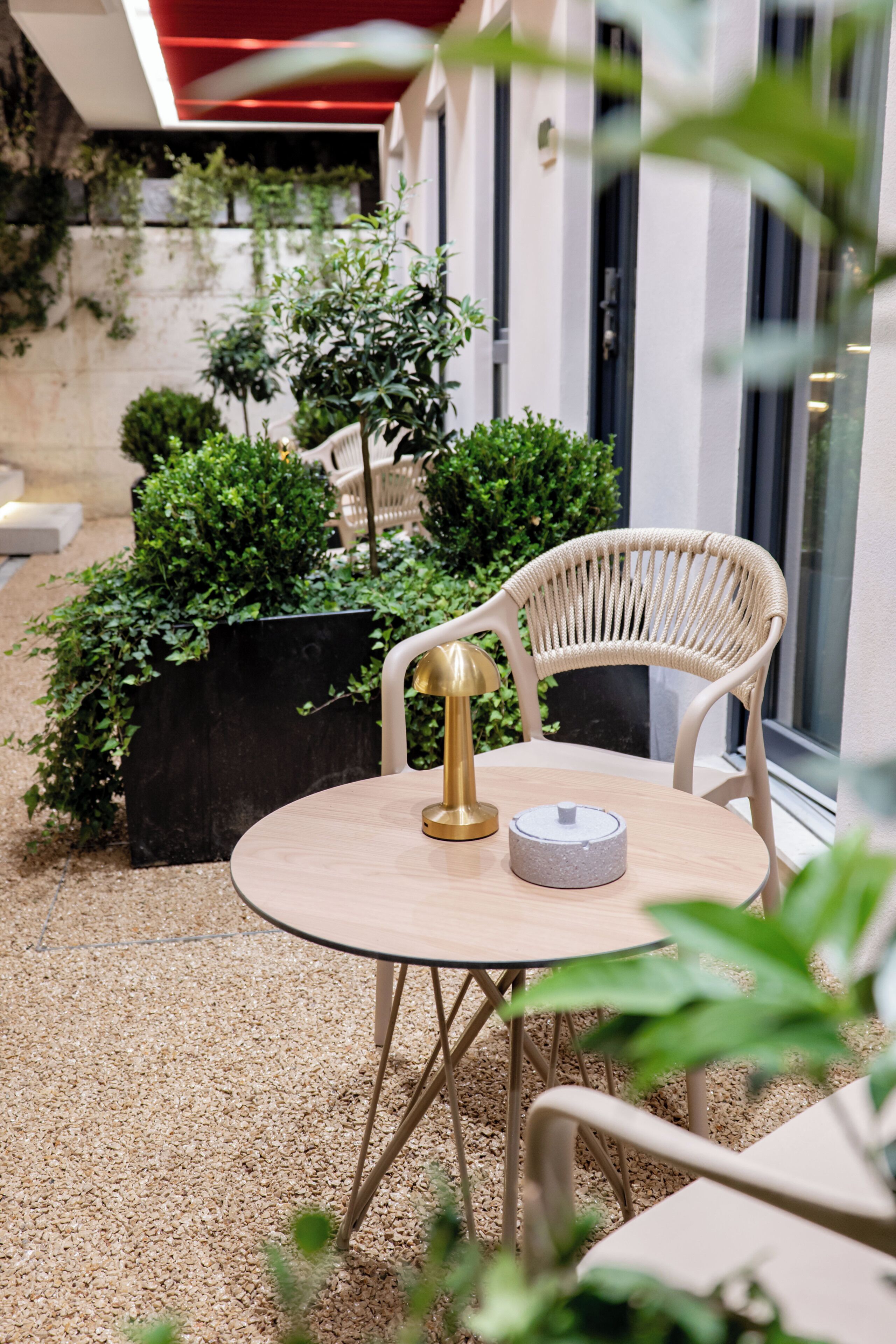 Une table ronde surmontée d’une lampe dorée, une chaise blanche en osier et un pot de fleurs avec des plantes vertes.