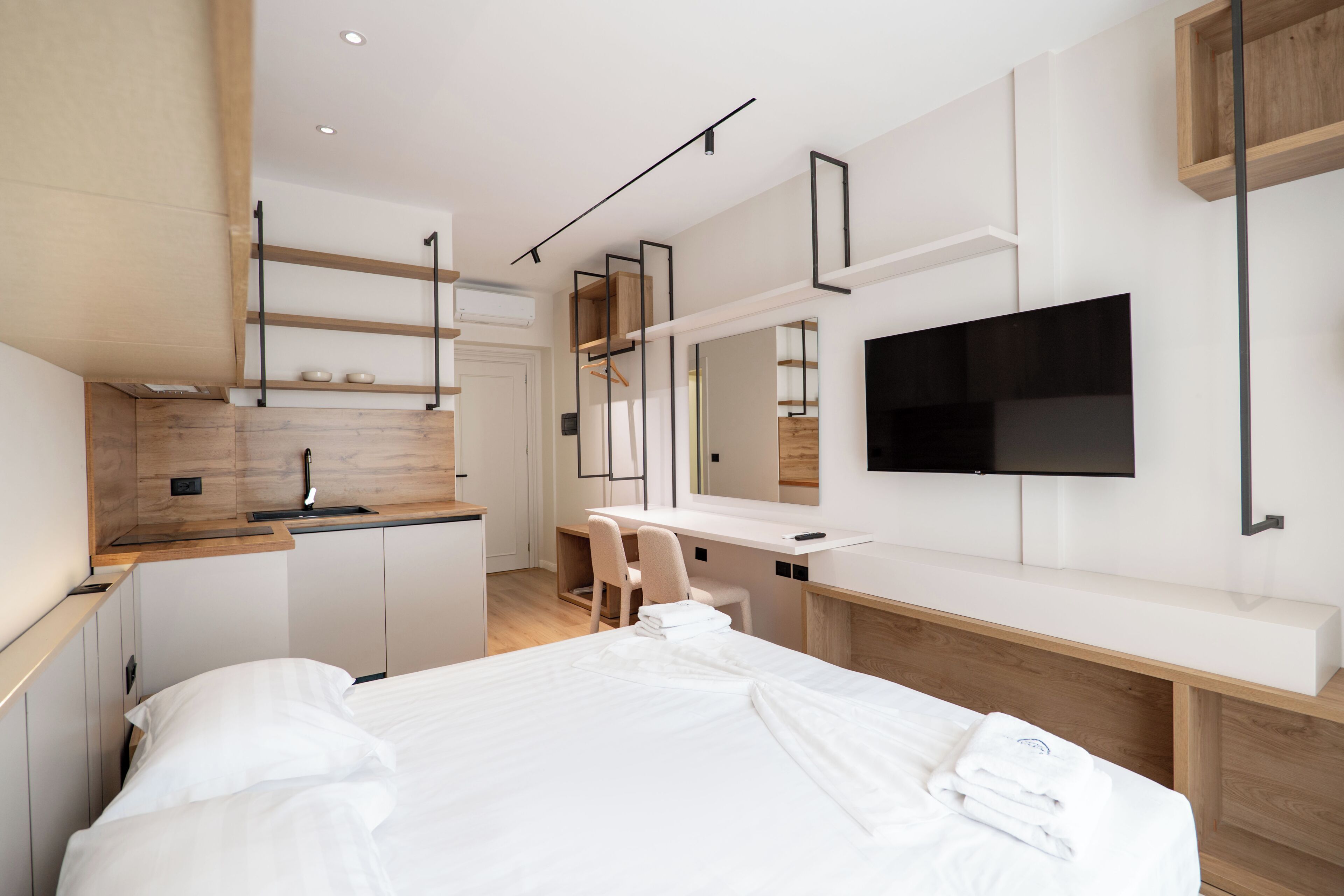 A modern hotel room with a bed, a desk, a chair, a TV, and a kitchenette.