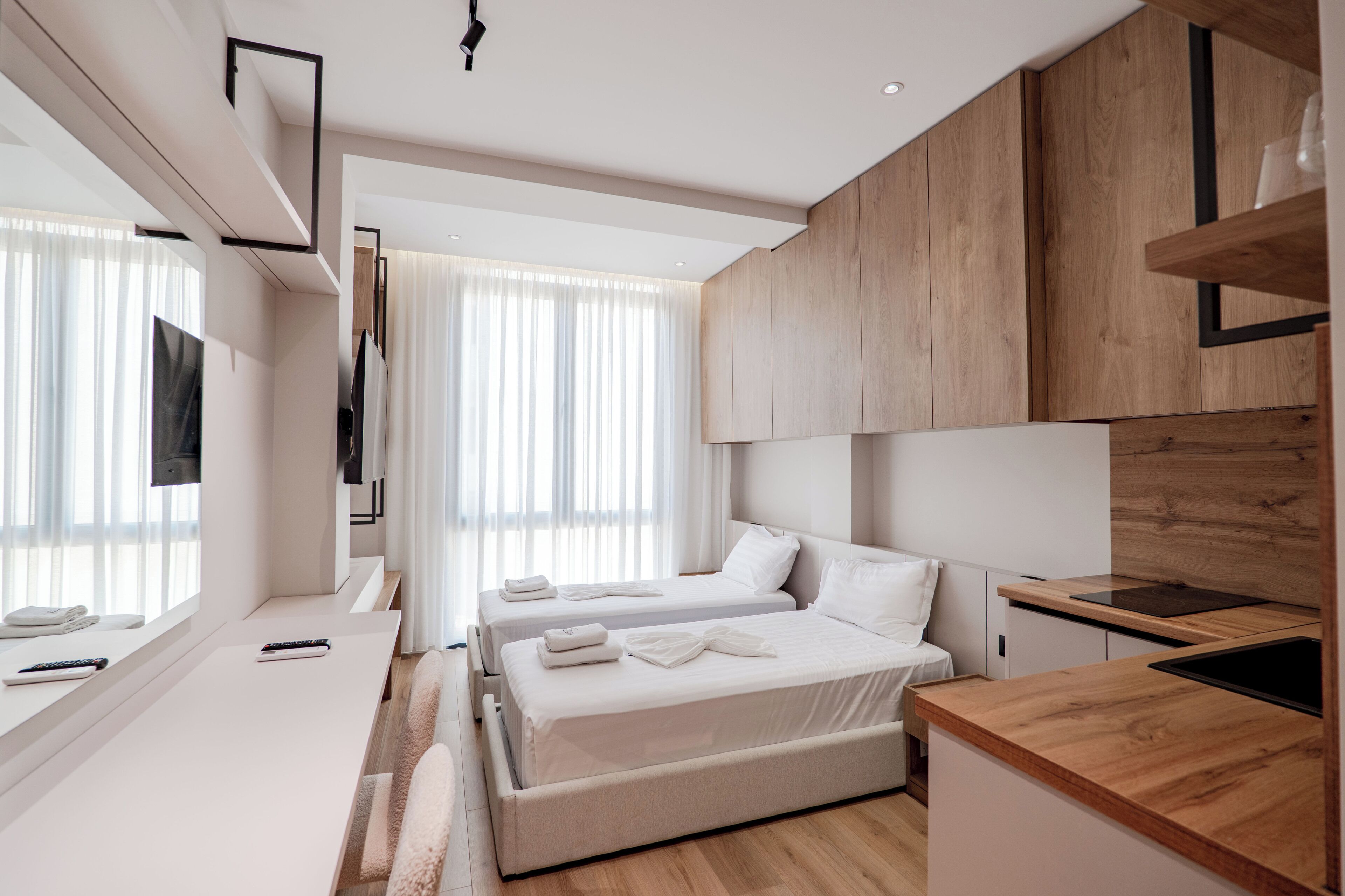 A modern hotel room with a large bed, a desk, a flat-screen TV, and a wooden kitchen area.