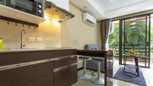 A modern kitchen with a microwave, stove, and sink, a bar area with stools, and a balcony with a view of palm trees. - MBC MaiKhao 1BR Condo•Poolside•Walk 2 Beach C194 (Tambon Mai Khao)