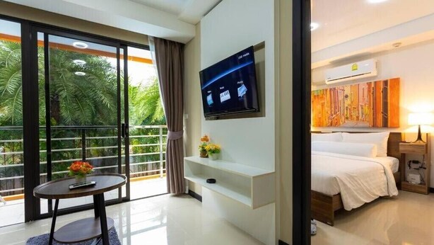 A hotel room with a bed, a TV mounted on the wall, a small table with a vase of flowers, and a balcony with a view of palm trees. - MBC MaiKhao 1BR Condo•Poolside•Walk 2 Beach C194 (Tambon Mai Khao)