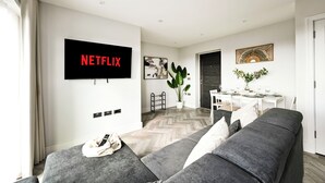 A modern living room with a grey sofa, a flat-screen TV mounted on the wall, a dining area with a table and chairs, and a potted plant. - 4-Guest North London | Piccadilly Line into Zone 1 (Barnet)