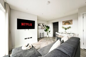 Ein modernes Wohnzimmer mit grauem Sofa, einem am Wand montierten Flachbildfernseher, einem Essbereich mit Tisch und Stühlen sowie einer Topfpflanze.