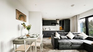 A modern kitchen with dark cabinets, a dining area with a white table and chairs, and a grey sofa with cushions. - 4-Guest North London | Piccadilly Line into Zone 1 (Barnet)