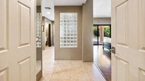 Un couloir avec un mur en blocs de verre, une porte sur la droite et une vue sur une chambre à coucher avec un lit et une fenêtre.