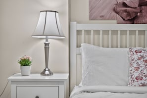A white bedside table with a lamp, a small potted plant, and a bed with white bedding and a floral pillow. - Tranquil Garden Level Retreat near UBC & Airport (Vancouver)