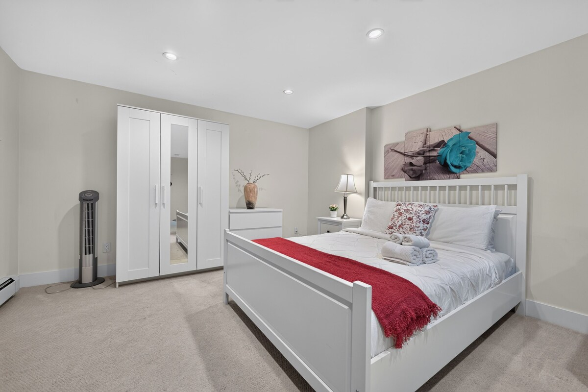 Un dormitorio con cama blanca, una manta roja, una mesita de noche con lámpara y una decoración de rosas en la pared.