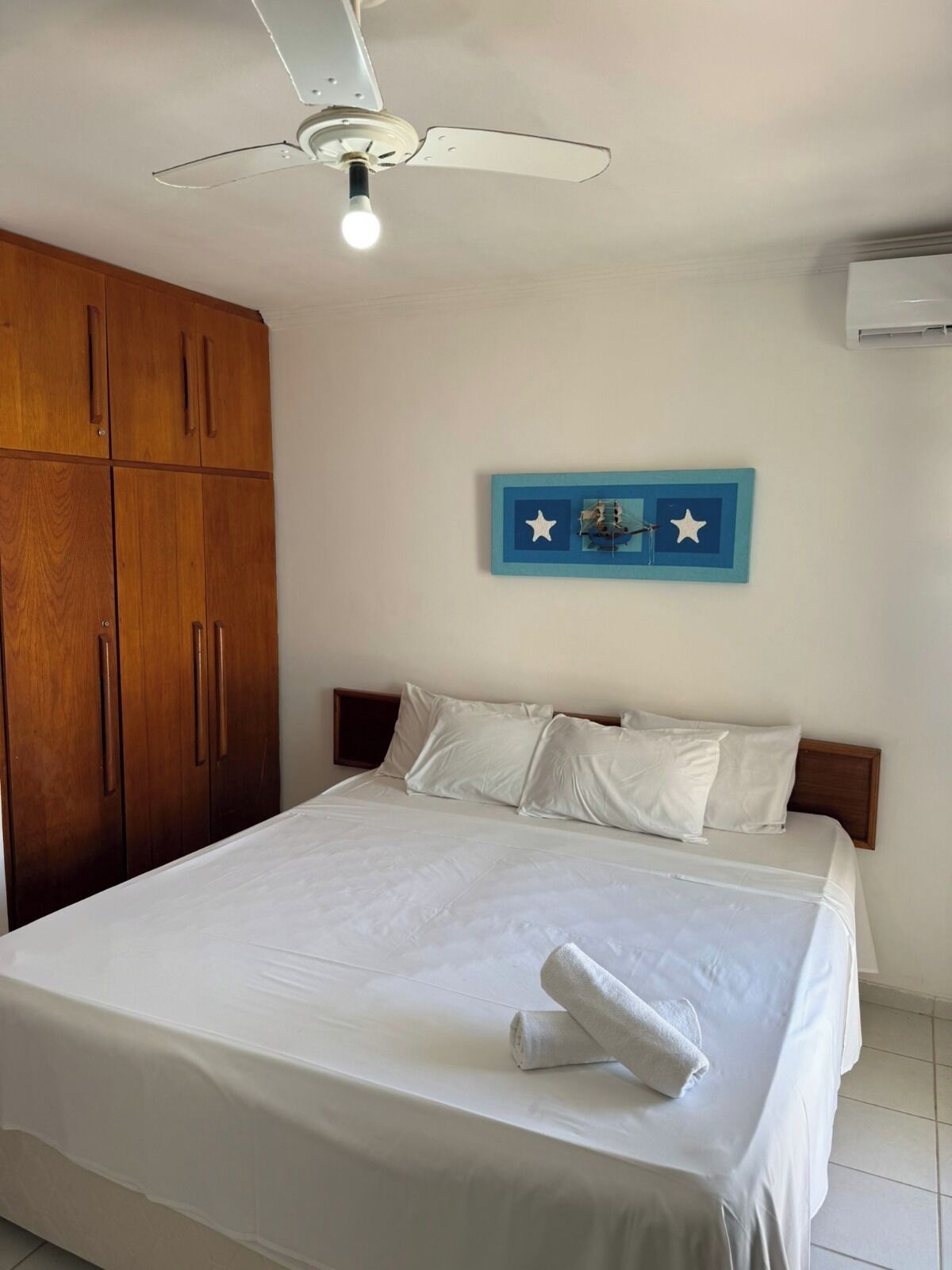 Habitación de hotel con cama, ropa de cama blanca, ventilador de techo, armario de madera y aire acondicionado de pared.