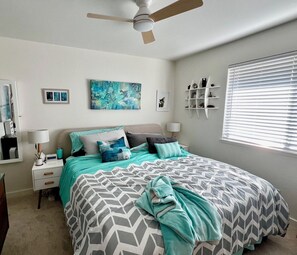 A bedroom with a bed, a ceiling fan, a nightstand, a lamp, a wall-mounted shelf, and a window with blinds.