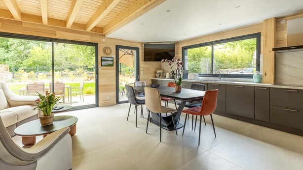 Cozinha moderna com grandes janelas, área de jantar com mesa e cadeiras, e sala de estar com sofá e mesa de centro.