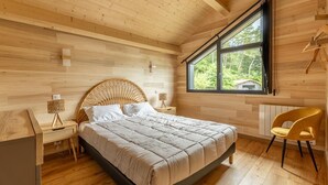 Um quarto com teto de madeira, uma janela grande, uma cama com cabeceira trançada, uma mesinha de cabeceira e uma cadeira.