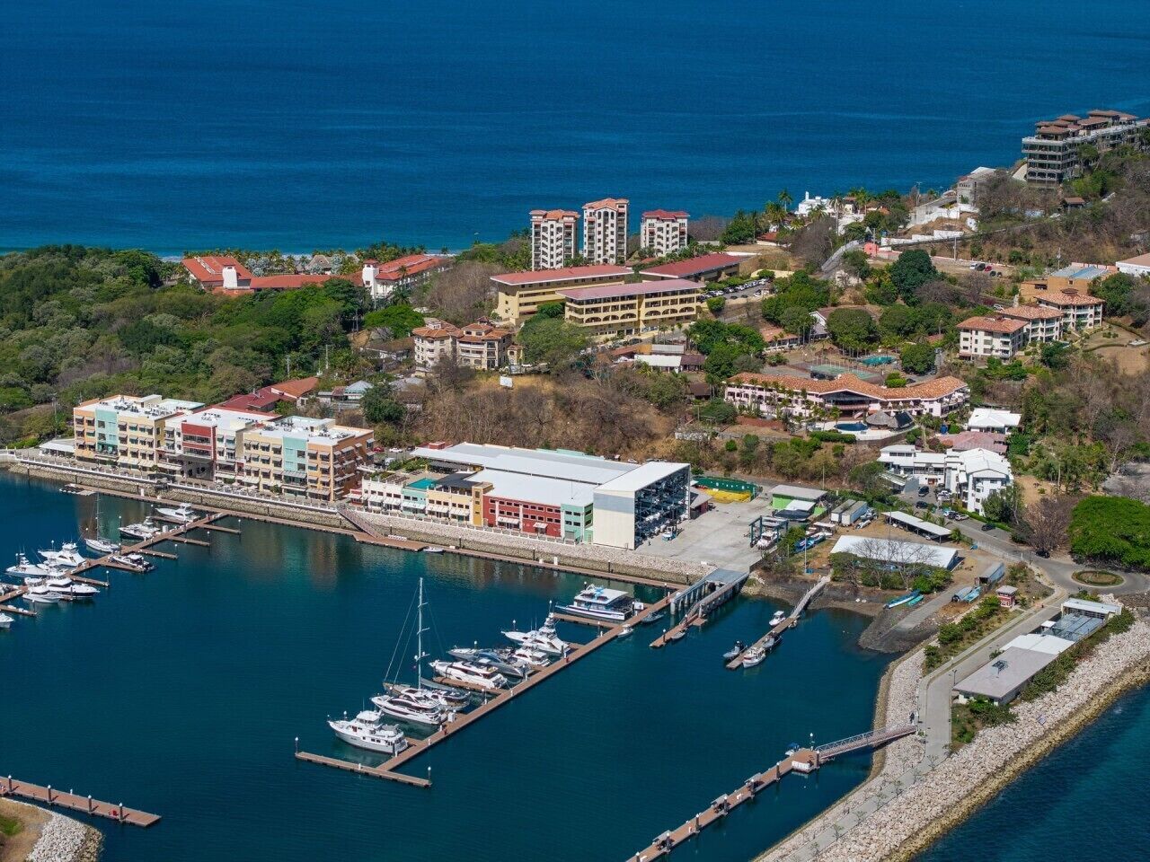 An aerial view of a coastal marina with multiple buildings, including a large multi-story structure and several smaller buildings, surrounded by water and greenery.