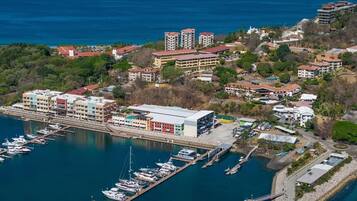 An aerial view of a coastal marina with multiple buildings, including a large multi-story structure and several smaller buildings, surrounded by water and greenery.