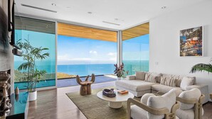A modern living room with a large sectional sofa, a coffee table, and a view of the ocean through floor-to-ceiling glass doors. - Casa Azure Estate by Oraklus (Malibu)