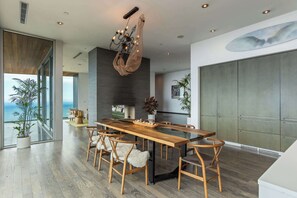 Une salle à manger moderne avec une table et des chaises en bois, un lustre et une grande fenêtre donnant sur l’océan.