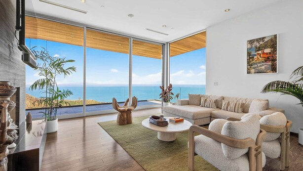 A modern living room with a large window overlooking the sea, a sofa, a coffee table, and a wooden accent wall. - Casa Azure Estate by Oraklus (Malibu)