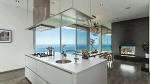 A modern kitchen with a large island, a dining area with a view, and a fireplace. - Casa Azure Estate by Oraklus (Malibu)