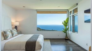 A bedroom with a large window, a bed, a nightstand, and a potted plant. - Casa Azure Estate by Oraklus (Malibu)