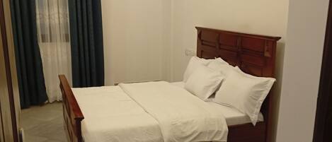 Un dormitorio con cama de madera, ropa de cama blanca, alfombra a rayas, una cortina y una lámpara fijada a la pared.