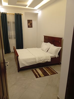 Un dormitorio con cama de madera, ropa de cama blanca, alfombra a rayas, una cortina y una lámpara fijada a la pared.