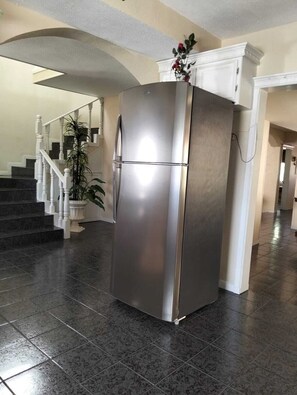 Un réfrigérateur en acier inoxydable se trouve dans un couloir, près d’un escalier et d’une plante en pot.