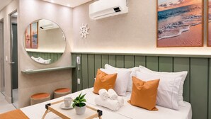 A modern hotel room with a bed, a bedside table with a tray, a mirror, a wall-mounted air conditioner, and a painting of a beach scene. - Nawi Beachfront Escape Apartment - Pool, Gym & Ocean Access (João Pessoa)