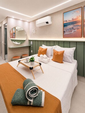Quarto de hotel moderno com uma cama, uma mesa de cabeceira com bandeja, um espelho, ar condicionado fixado na parede e uma pintura de uma cena de praia.