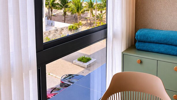 A room with a view of a beach, a table with a wooden surface, a chair, and a green cabinet with a towel. - Nawi Beachfront Escape Apartment - Pool, Gym & Ocean Access (João Pessoa)