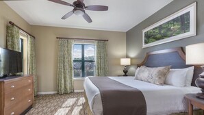 Una habitación de hotel con una cama, un televisor sobre una cómoda, un ventilador de techo, una ventana con cortinas, un cuadro en la pared y una lámpara en una mesita de noche.