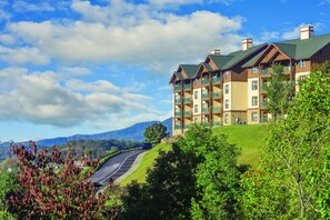 Un hotel de varios pisos con balcones, situado en una ladera con vistas a montañas y una carretera sinuosa.