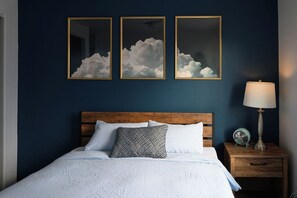 Ein Schlafzimmer mit Holz-Kopfteil, einem Bett mit weißer Bettwäsche, drei gerahmten Gemälden mit Wolkenmotiven an der Wand, einer Nachttischlampe und einem kleinen Ventilator auf dem Nachttisch.