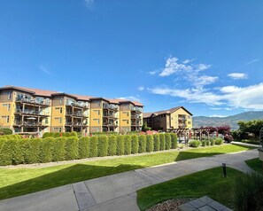 A multi-story residential building with balconies, a well-maintained lawn, and a clear view of a mountain range in the background. - Barona Beach lakefront Resort -Main floor condo - Pool-Hot tub-Private beach (West Kelowna)