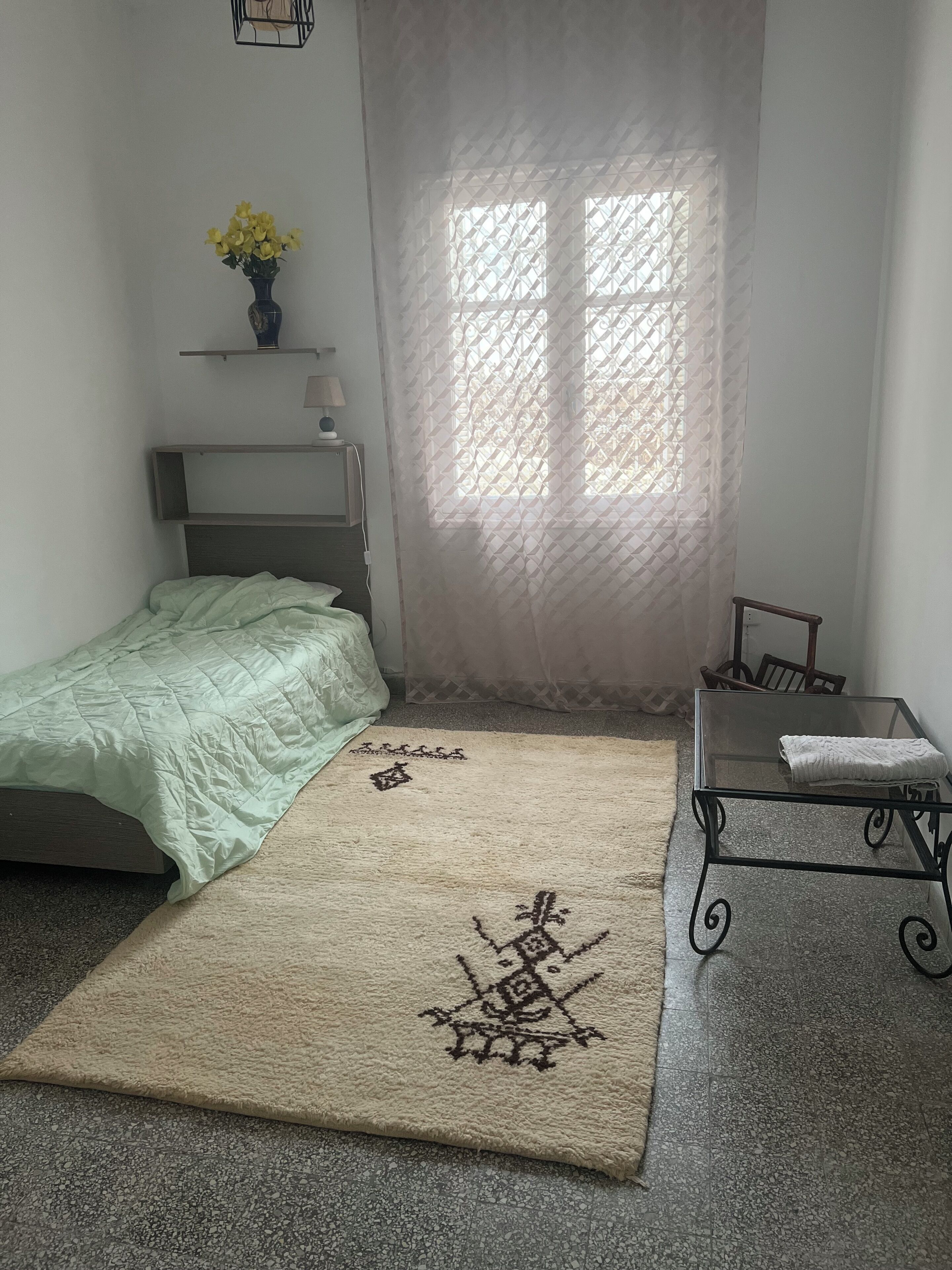 Un dormitorio con cama, una mesita, una silla, un ventanal con cortina estampada y una alfombra decorativa.