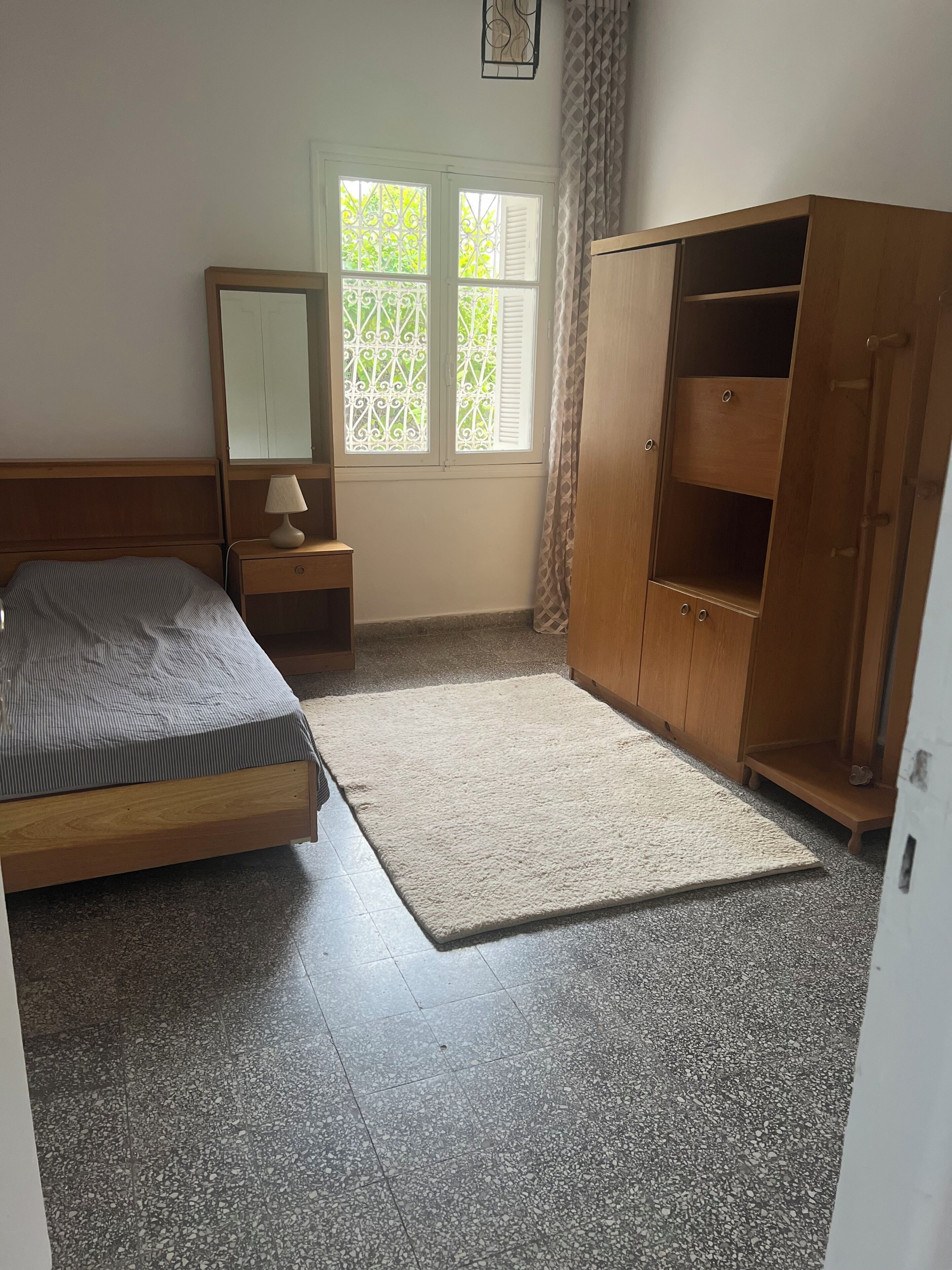 Een slaapkamer met een bed, nachtkastje, dressoir, raam met witte luiken en uitzicht op groen.