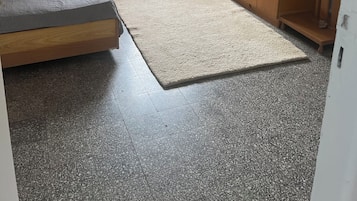 Een slaapkamer met een bed, nachtkastje, dressoir, raam met witte luiken en uitzicht op groen.