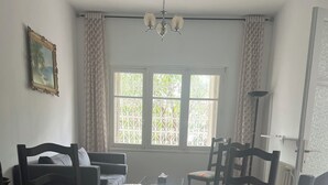 A dining area with a glass-top table surrounded by chairs, a chandelier, and a window with decorative iron grilles.