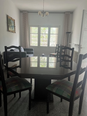 A dining area with a glass-top table surrounded by chairs, a chandelier, and a window with decorative iron grilles.