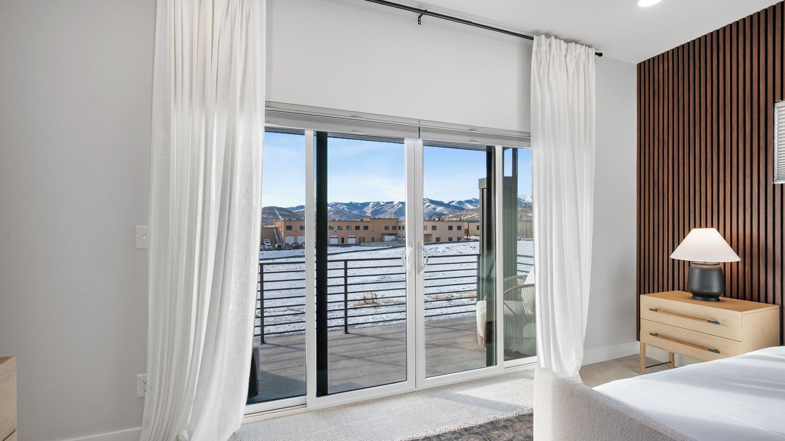 A modern bedroom with a large sliding glass door to a balcony, a bedside table with a lamp, and a view of buildings and mountains.