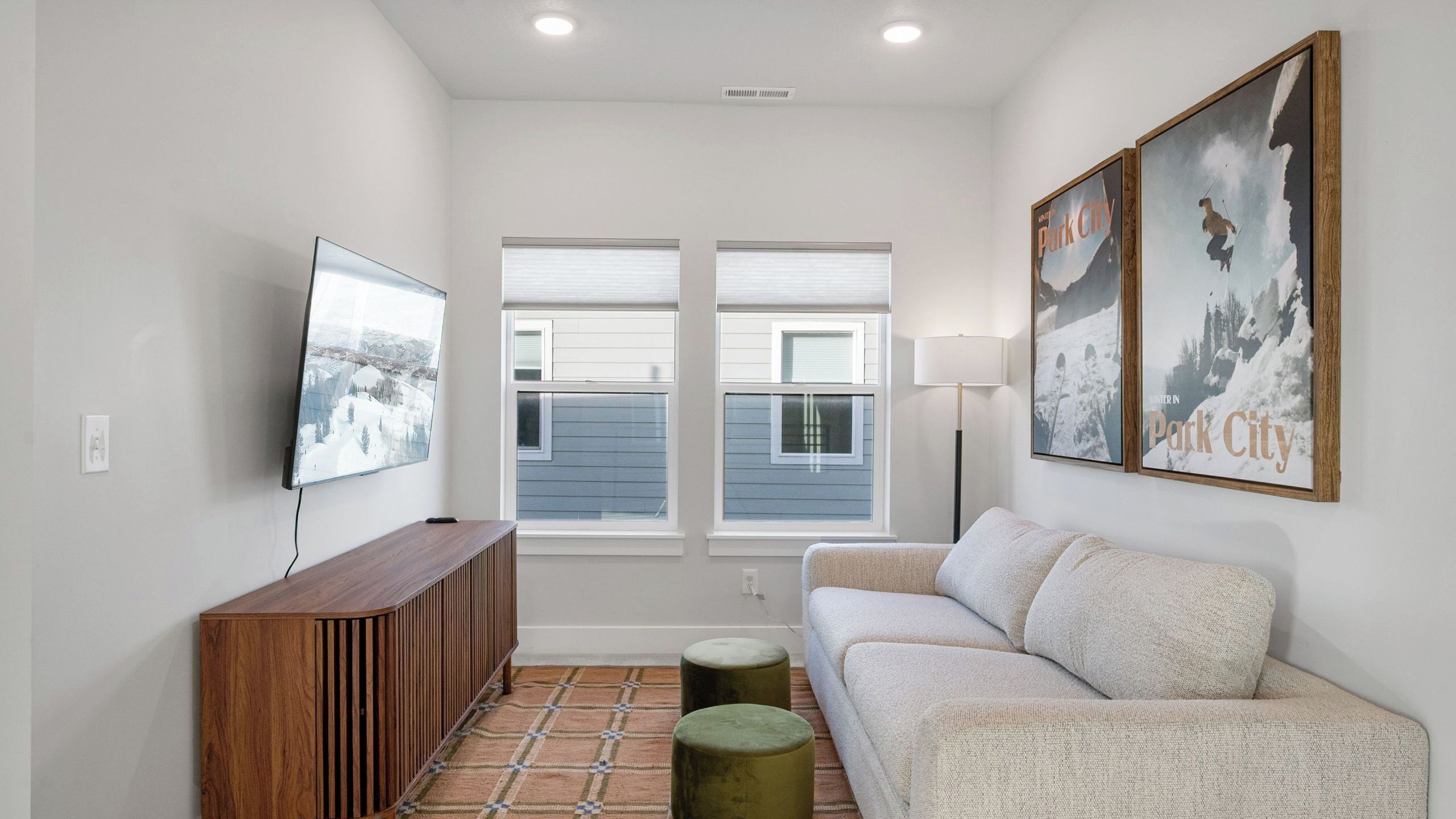A modern living room with a flat-screen TV, a wooden console, a light-colored sofa, two round ottomans, and a large framed picture of a snowy mountain.