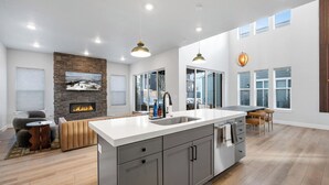 A spacious kitchen with a large island, a stone fireplace, and a view through the glass doors. - Poppy & Pine by Avantstay Sleeps 14 + Hot Tub Game Room + Fire Pit Silver Creek + Canyons (Park City)
