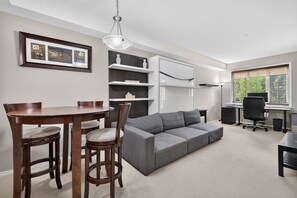 A modern living space with a dining table, chairs, a gray sofa, a desk with a computer, and a window with blinds. - Mountain View Condo | Pool, Hot Tub & Golf 2213 (Kelowna)