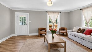 Ein Wohnzimmer mit Holzboden, einer weißen Tür, zwei Ledersesseln, einem beigefarbenen Sofa, einem Holztisch und einer Vase mit Blumen.