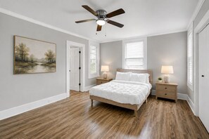 Une chambre à coucher avec un lit, deux tables de chevet avec des lampes, un ventilateur de plafond, un tableau au mur et une porte donnant sur une autre pièce.