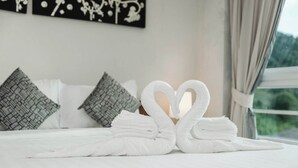 Una cama con sábanas blancas, dos toallas con forma de corazón, almohadas estampadas y una ventana con vistas.
