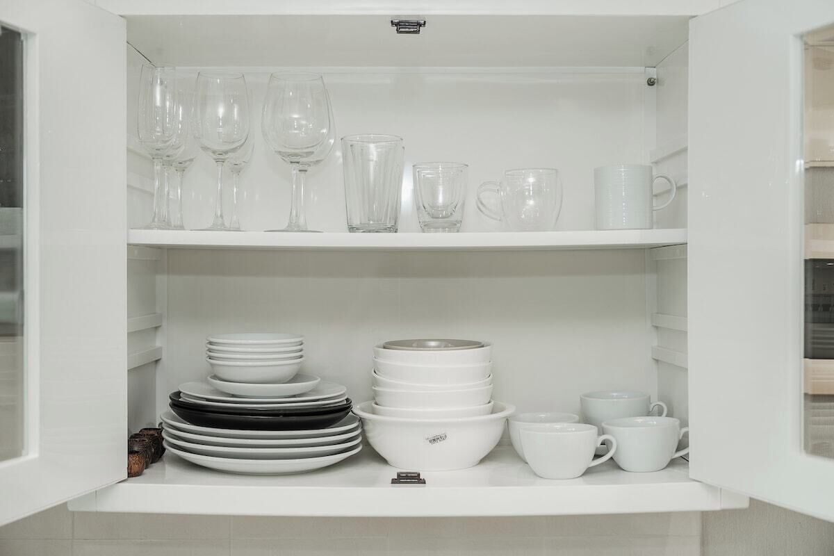 Un armario blanco con estantes que contienen copas de vino transparentes, tazas blancas y platos apilados.