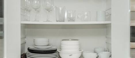 Un armario blanco con estantes que contienen copas de vino transparentes, tazas blancas y platos apilados.